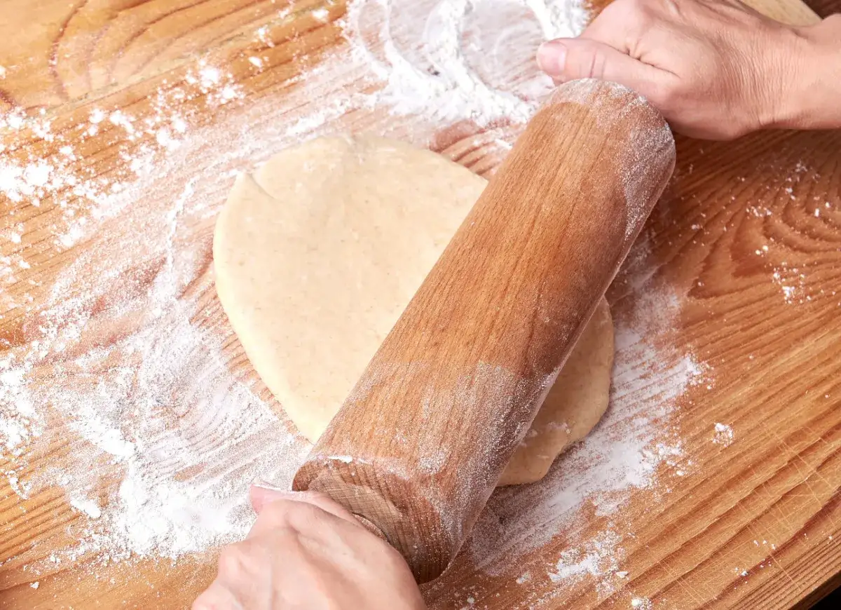 Ręce wałkują ciasto na pierogi na drewnianej desce. Mąka rozsypana wokół.