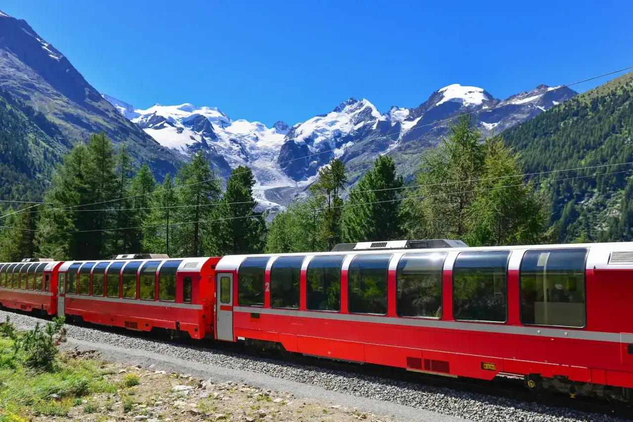 Treno rosso panoramico sulla ferrovia del Bernina, con maestose montagne innevate sullo sfondo e fitti boschi di conifere.