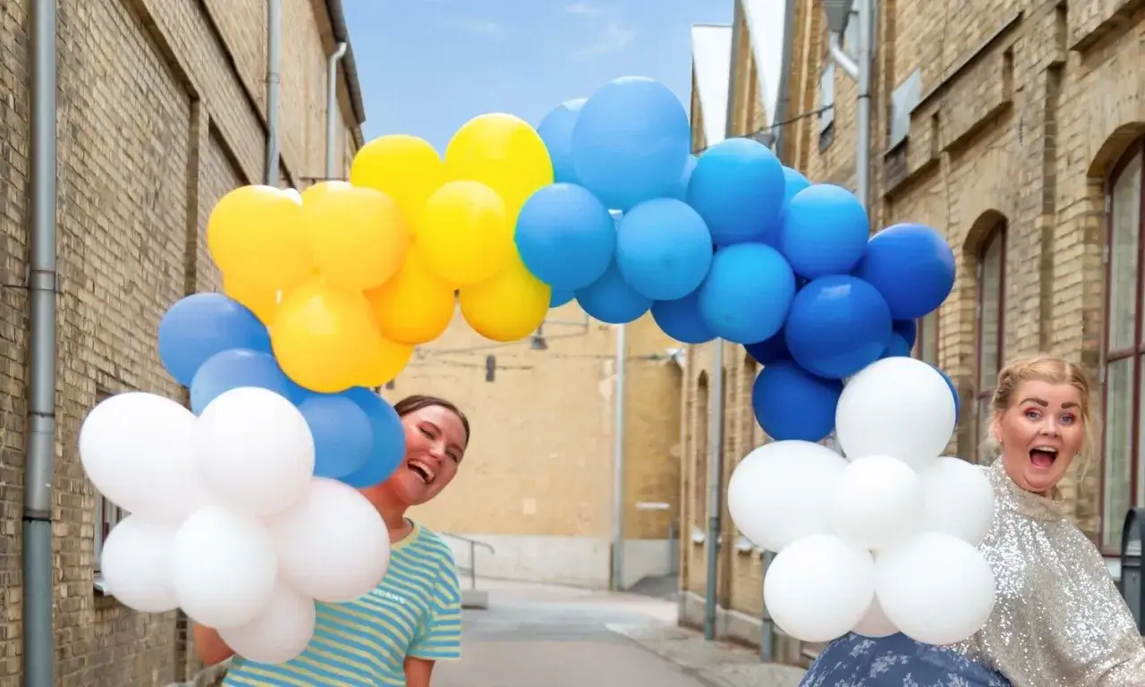 Tv&aring; glada kvinnor firar en studentfest med en b&aring;ge av bl&aring;, gula och vita ballonger.