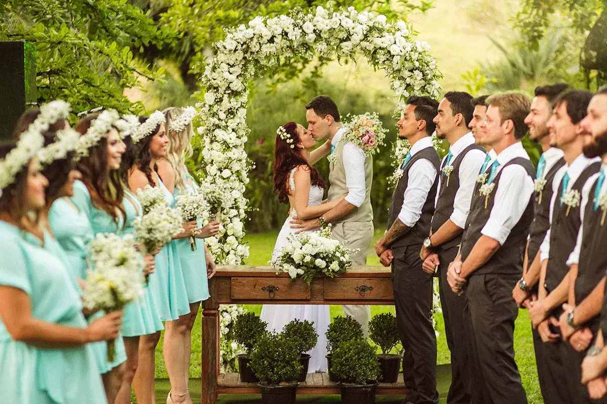 La pareja se besa bajo un arco de flores. A su lado, la dama de honor y el caballero de honor observan la escena.