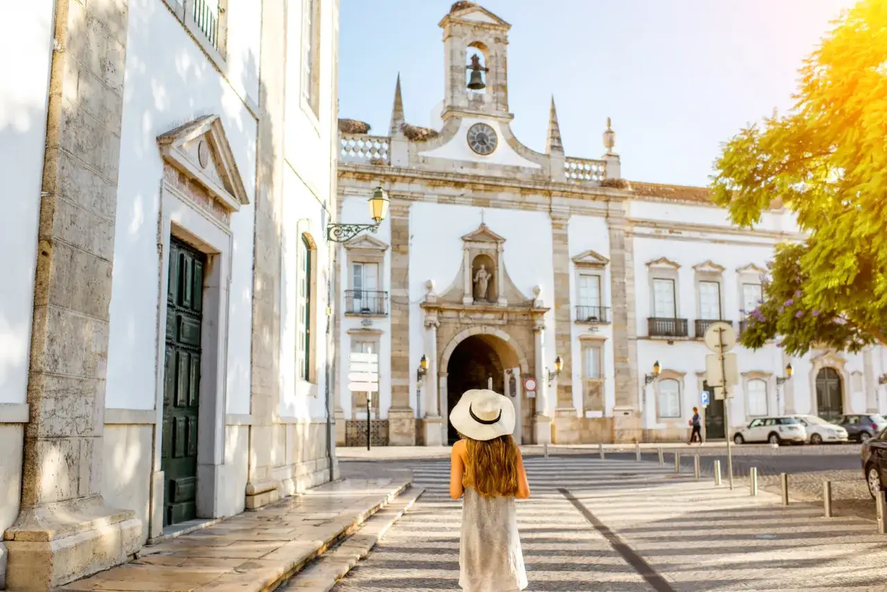 Frau mit Hut steht vor historischem Gebäude in Faro, Portugal. Sonnenschein beleuchtet die Architektur.