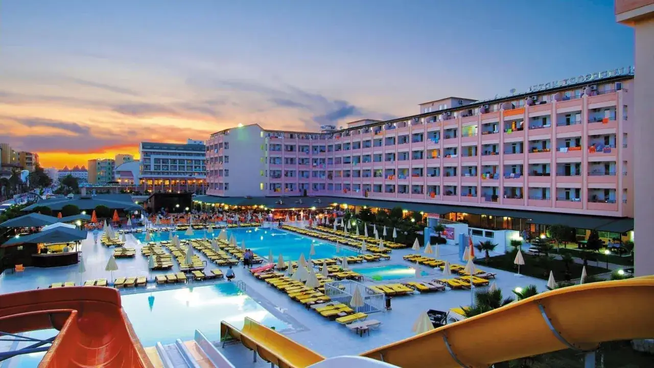 Xeno Eftalia Resort: rozległy kompleks basen&oacute;w z leżakami i zjeżdżalniami wodnymi, otoczony hotelami i palmami, o zachodzie słońca.