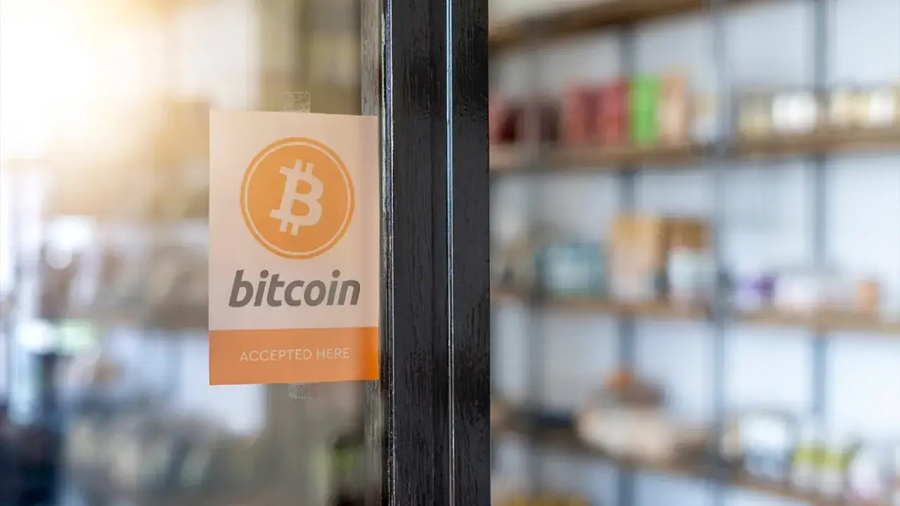 Naklejka na drzwiach informuje, gdzie można płacić bitcoinem. W tle widać p&oacute;łki sklepowe.