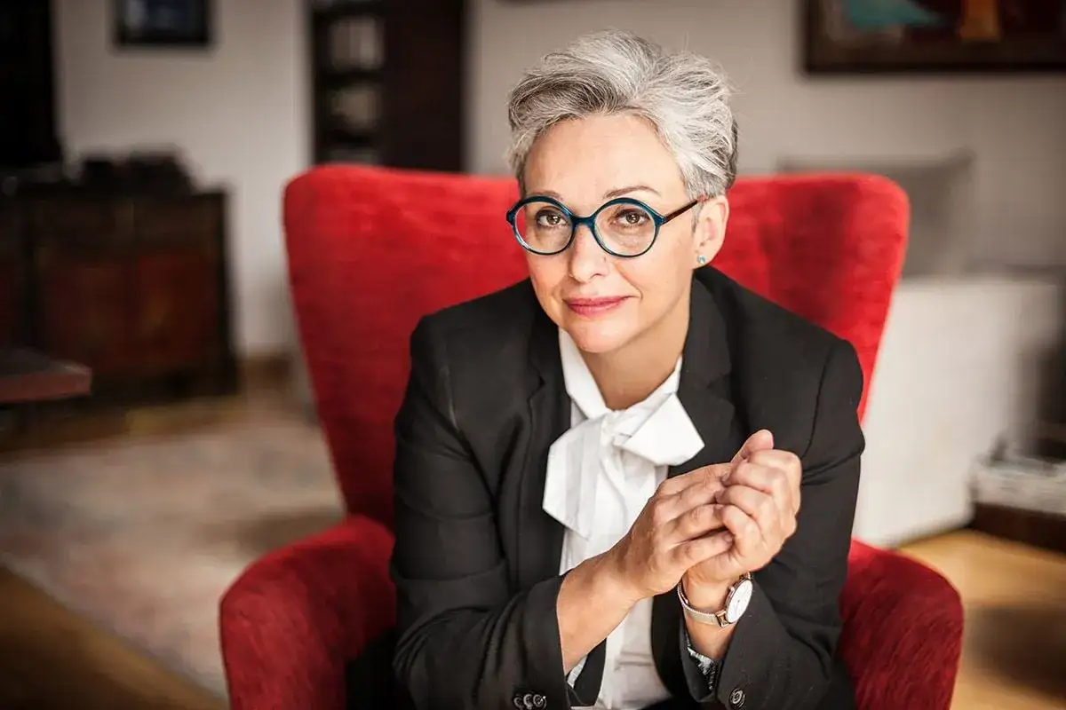 Kinga Dębska w okularach i czarnym żakiecie siedzi w czerwonym fotelu, z dłońmi złożonymi na kolanach.