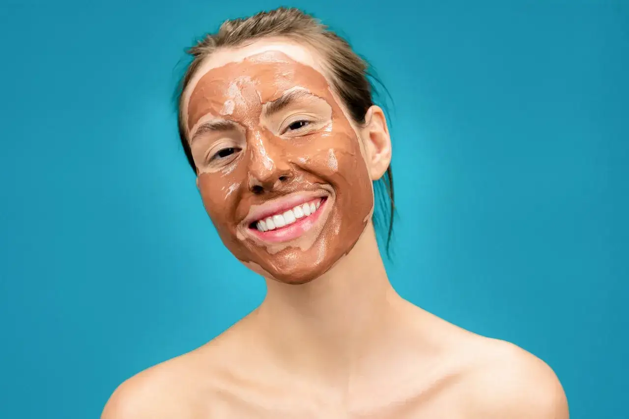 Ragazza sorridente con una maschera all'argilla sul viso, che promette un viso lucido e radioso.