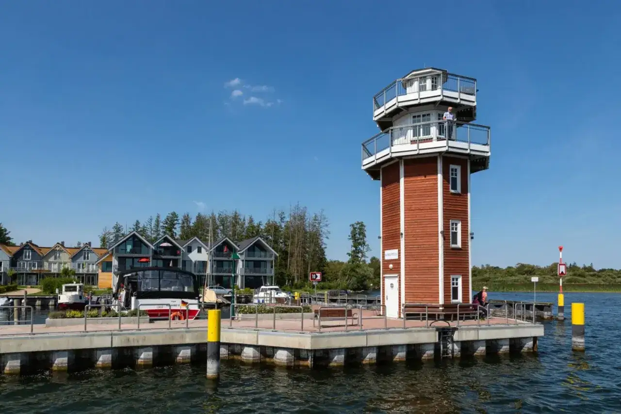 Ein Mann steht auf dem Aussichtsdeck des roten Leuchtturms. Im Hintergrund reihen sich moderne Häuser am Wasser. Ein schöner Tag am See mit Promenade.