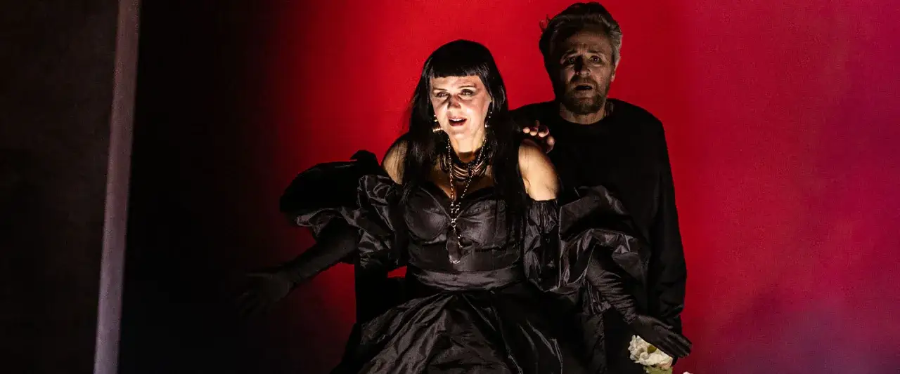 Dramatyczna scena z opery Karola Szymanowskiego: kobieta w czarnej sukni i mężczyzna z wyrazem przerażenia na twarzy na tle czerwonej kurtyny.
