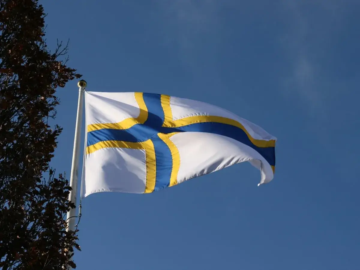 Sverigefinnarnas dag firas med en vit flagga med blått och gult kors som vajar i vinden mot en klarblå himmel.