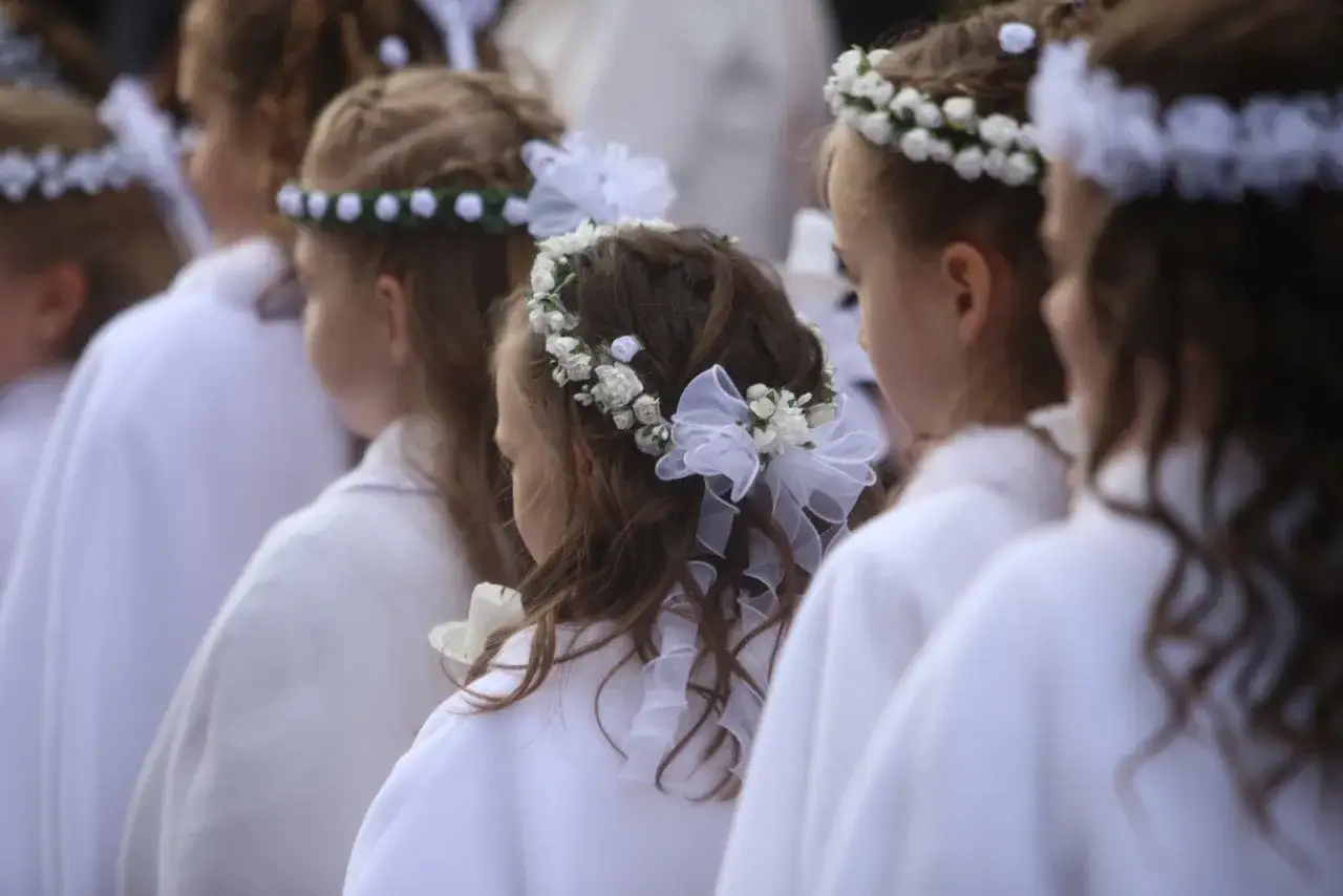 Urocza fryzura na komunię dla dziewczynki z wianuszkiem z białych kwiatów i kokardką.