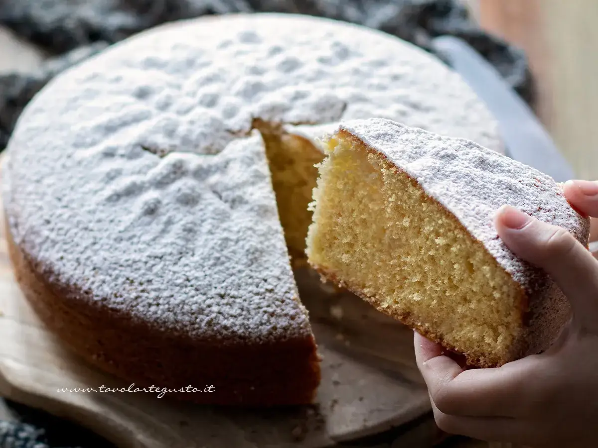 Una fetta di torta semplice, soffice e cosparsa di zucchero a velo, viene sollevata da una mano.