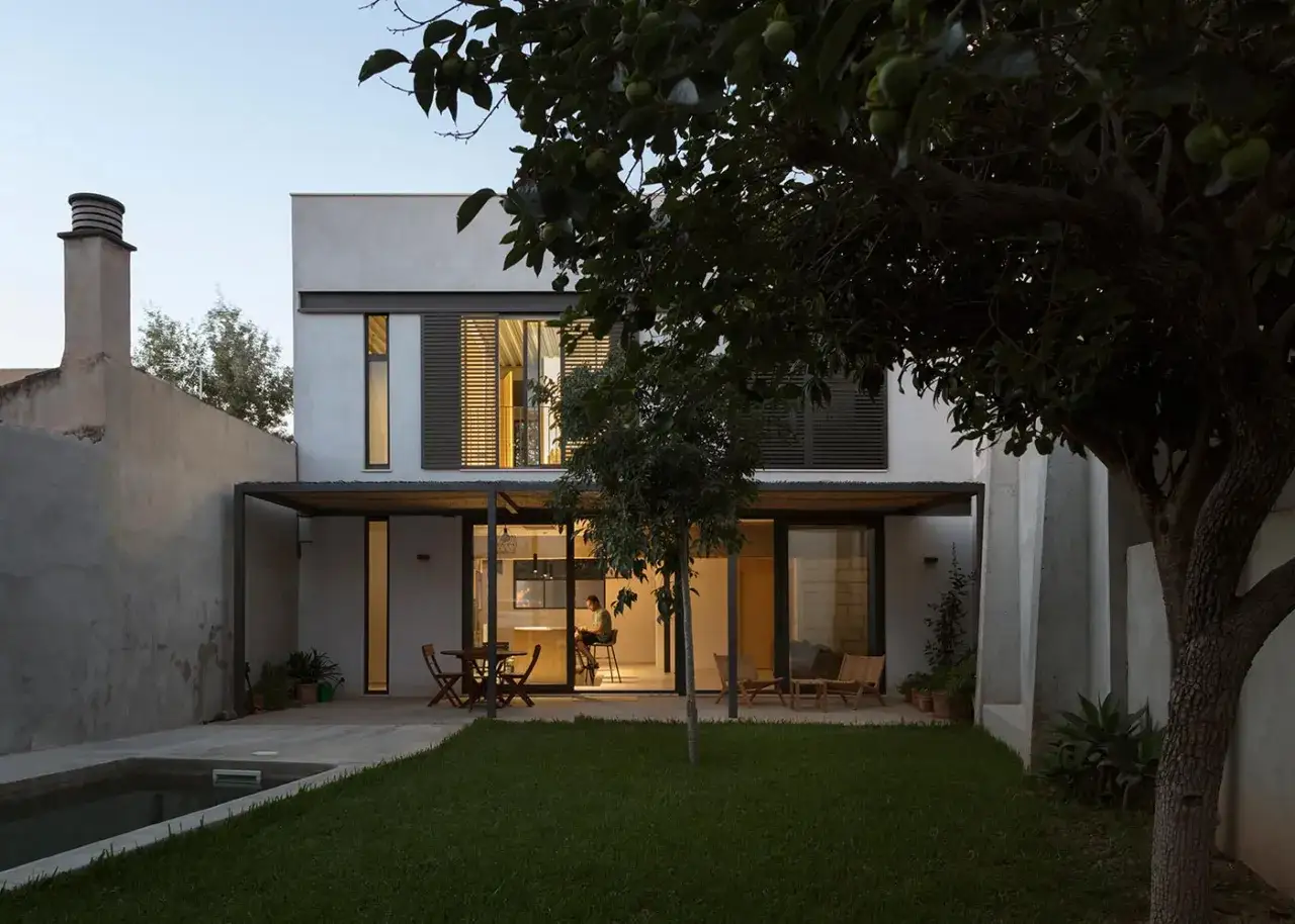 Moderna casa entre medianeras con piscina y jardín. La luz cálida del interior ilumina la cocina y el salón.
