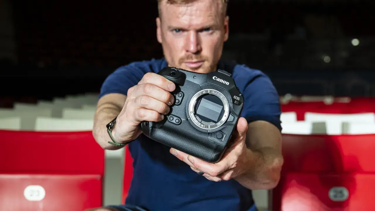 Mężczyzna prezentuje aparat Canon EOS R1, siedząc na czerwonych trybunach.