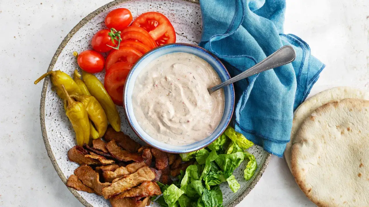 En skål med krämig kebabsås recept, bredvid skivad tomat, inlagda paprikor, sallad, kött och pitabröd.
