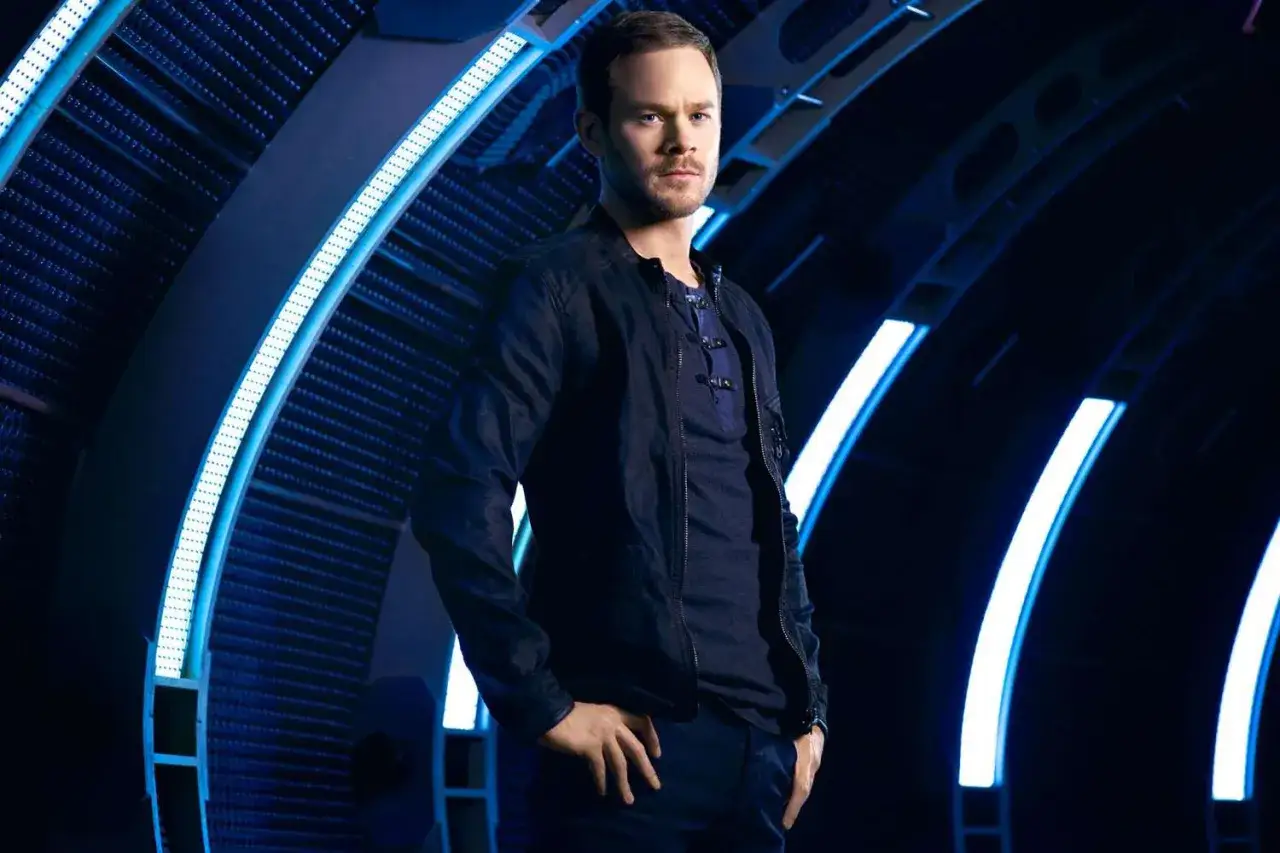 Aaron Ashmore, bekannt aus Filmen und Serien, steht in einem dunklen, futuristischen Korridor mit blauen Lichtern.