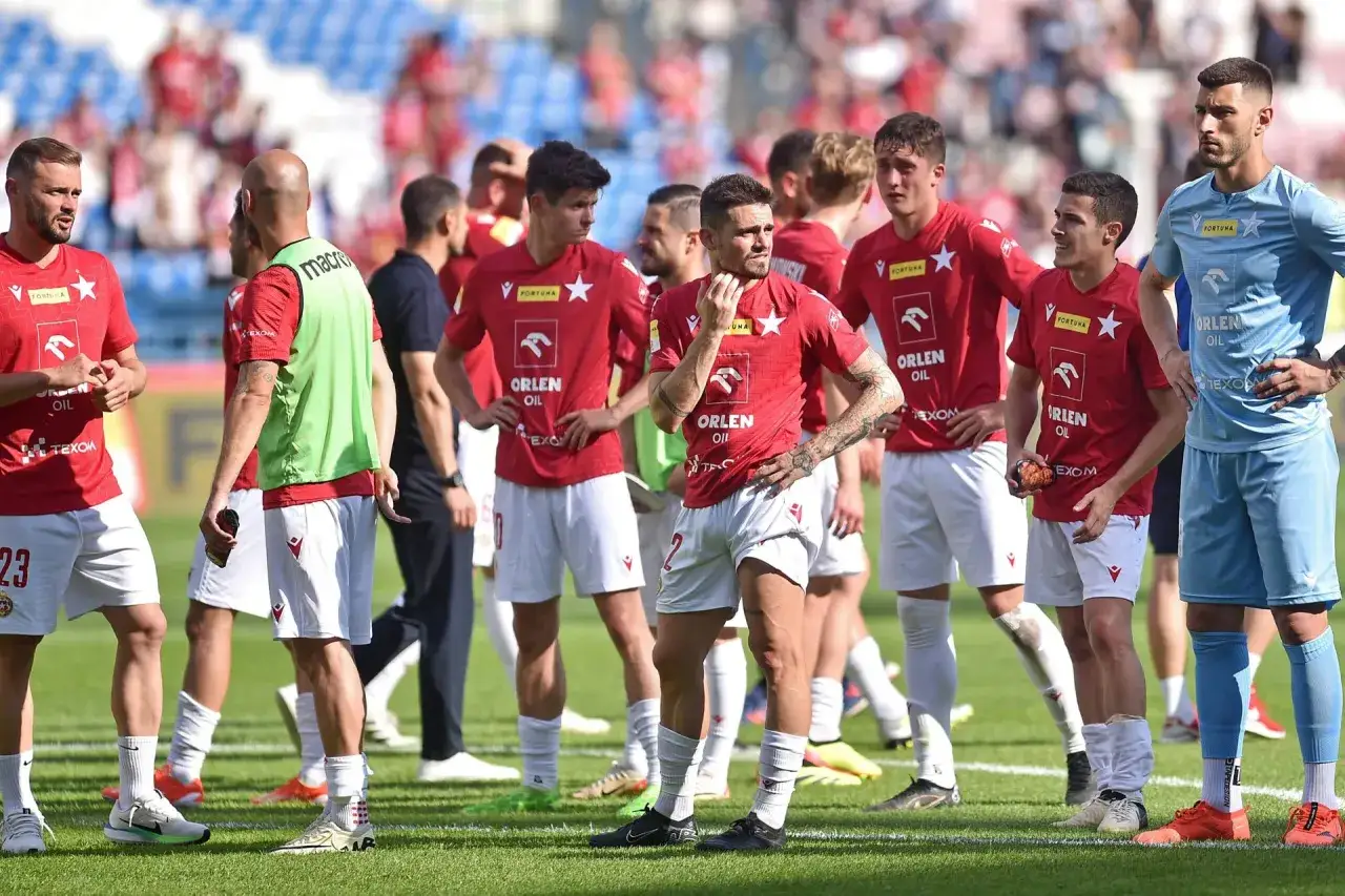 Piłkarze w czerwonych strojach, być może najgorszy klub na świecie, stoją na boisku. Jeden z nich ma zieloną kamizelkę.