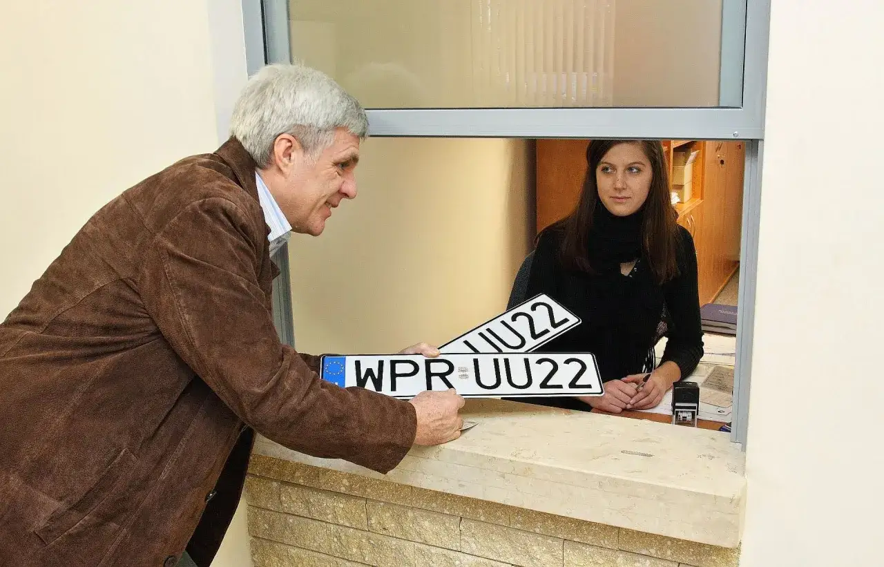 Mężczyzna z tablicami rejestracyjnymi pyta, czy trzeba zmieniać tablice rejestracyjne. Urzędniczka w okienku słucha.