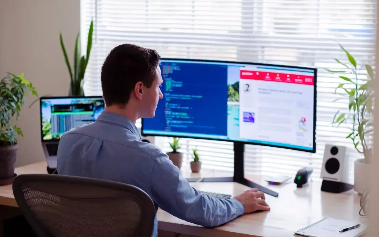 Uomo con buona postura pc, concentrato sul codice su un monitor curvo. Piante verdi e luce naturale completano l'ambiente di lavoro.