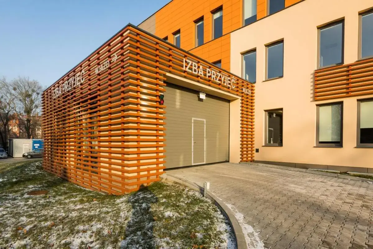 Nowoczesny budynek z pomarańczową fasadą i drewnianą ażurową zabudową przy wjeździe.