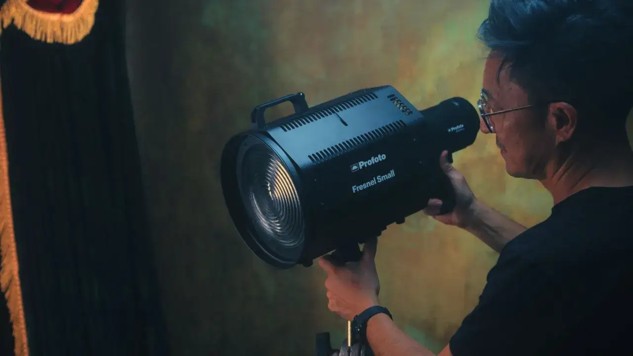 Mężczyzna z niebieskimi włosami i okularami trzyma lampę Profoto Fresnel Small, ustawiając światło.