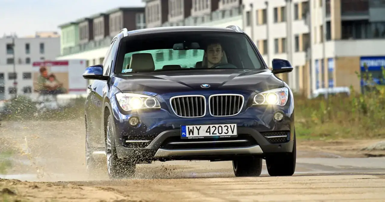 Ciemnoniebieskie BMW X1 przejeżdża przez kałużę, rozpryskując wodę. Kierowca zastanawia się, ile kosztuje BMW X1, planując zakup.
