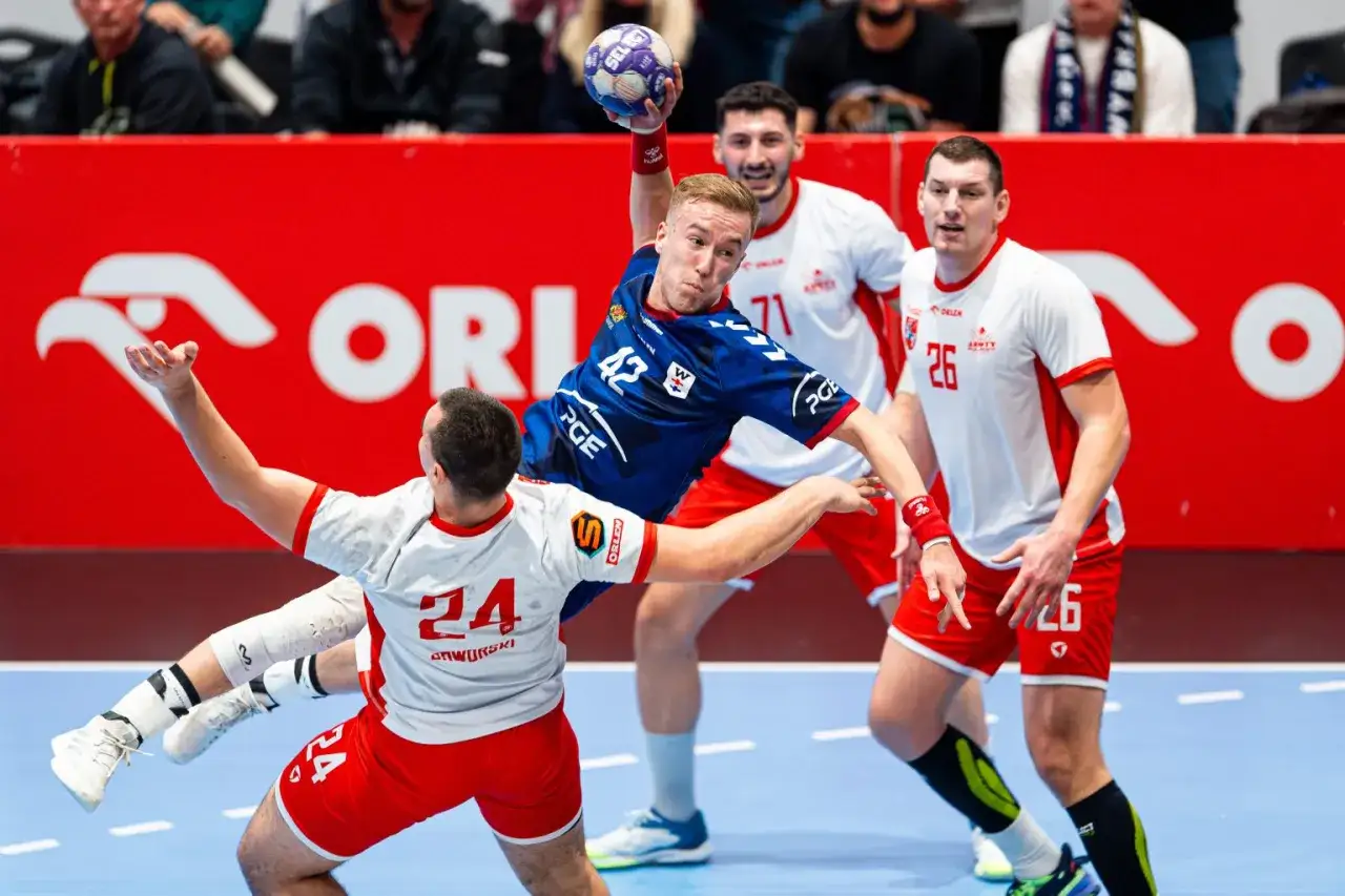 Piłkarz ręczny w skoku rzuca do bramki, broniony przez zawodnik&oacute;w w białych strojach z numerami 24 i 26. W tle reklama ORLEN.