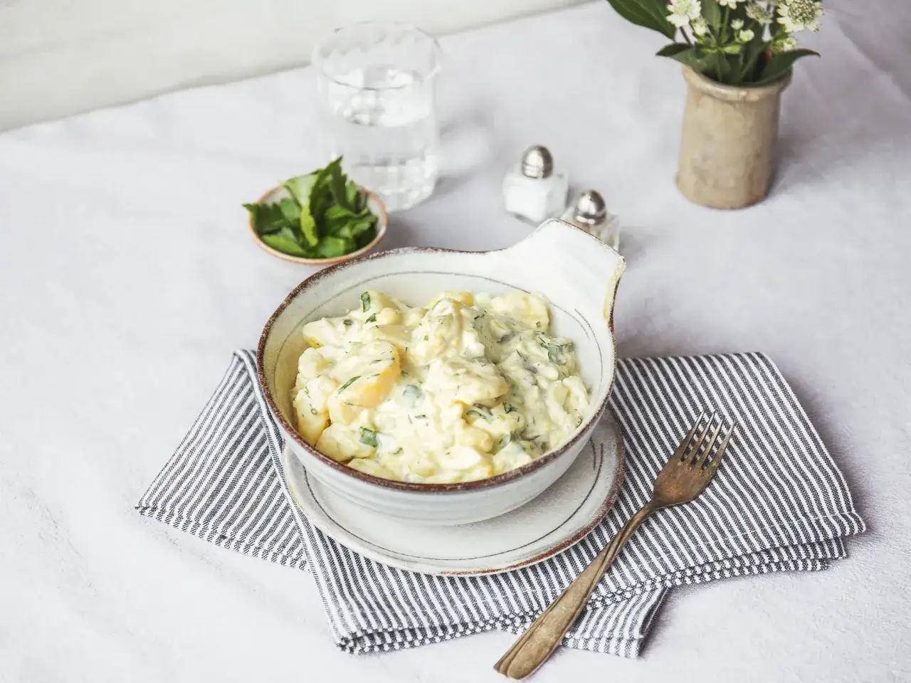 Ein cremiger Kartoffelsalat, der an Olivier Salat erinnert, serviert in einer Sch&uuml;ssel mit Kr&auml;utern und einem Glas Wasser.