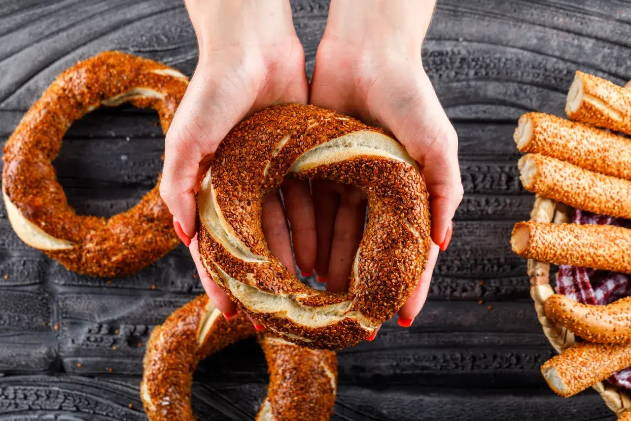 Le mani tengono un simit, un classico street food da provare a Istanbul. Sullo sfondo, altri simit e grissini ricoperti di sesamo.
