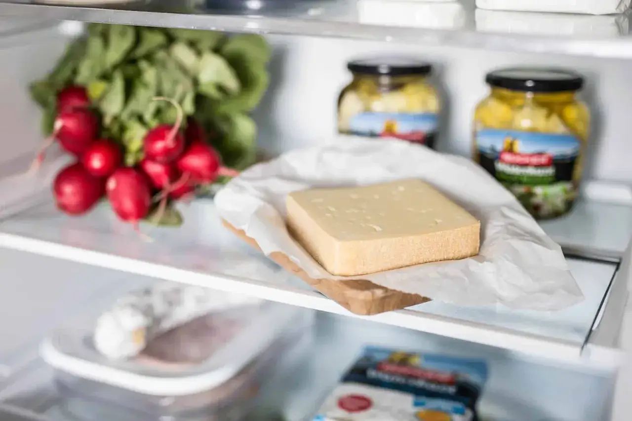Ein St&uuml;ck K&auml;se liegt auf einem Holzbrettchen mit Papier im K&uuml;hlschrank. So l&auml;sst sich K&auml;se, wie z.B. Feta, aufbewahren.