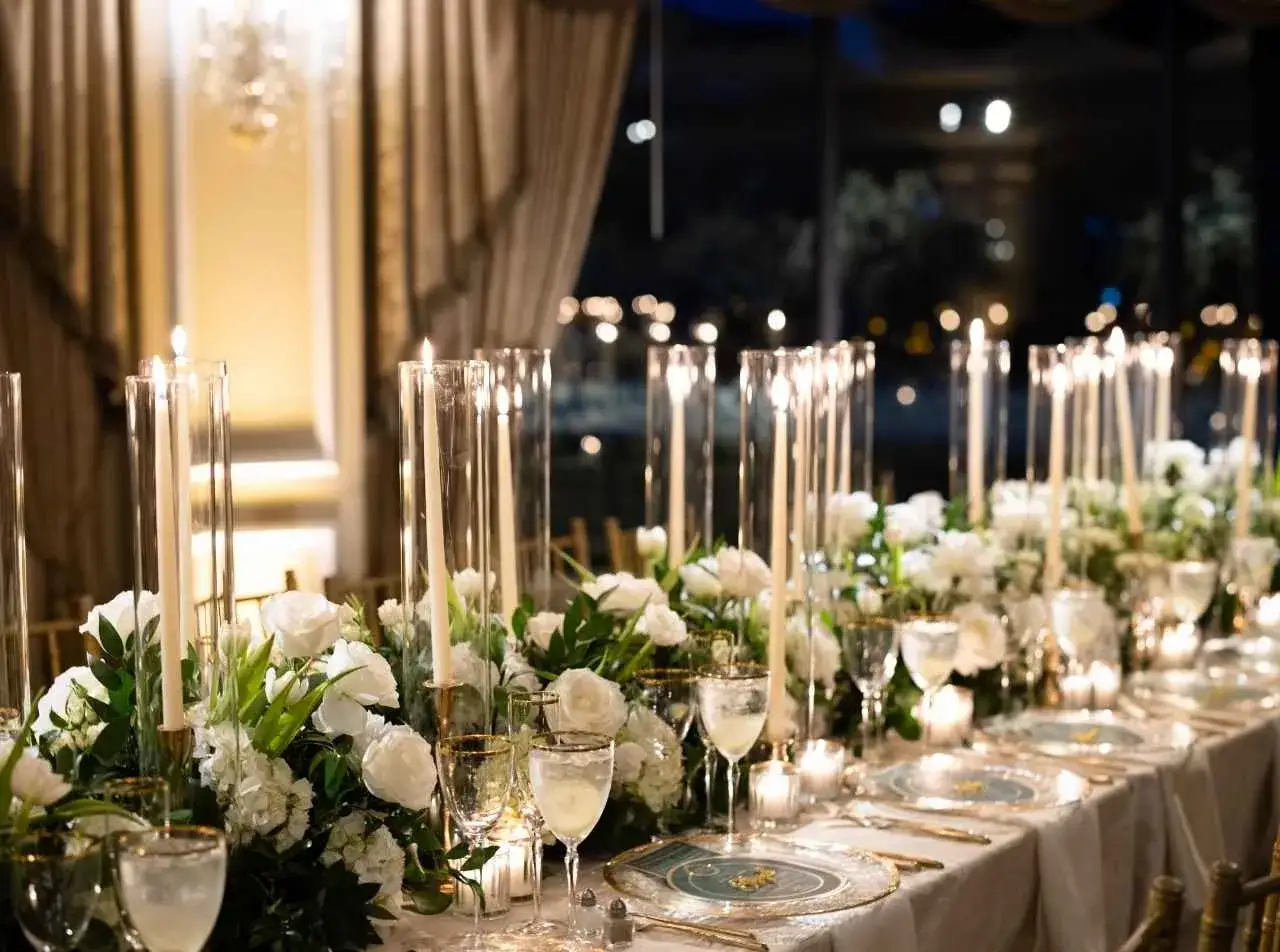 Elegancka sala weselna z długim stołem, udekorowanym białymi kwiatami, świecami w szklanych wazonach i złotymi sztućcami. Zastanawiasz się, ile kosztują kwiaty na wesele?