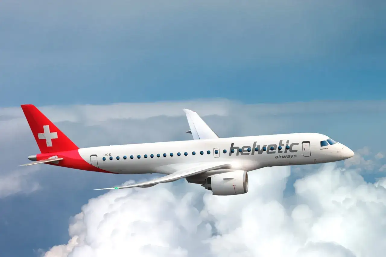 Samolot linii Helvetic Airways z charakterystycznym czerwonym ogonem i białym krzyżem leci na tle błękitnego nieba i chmur.