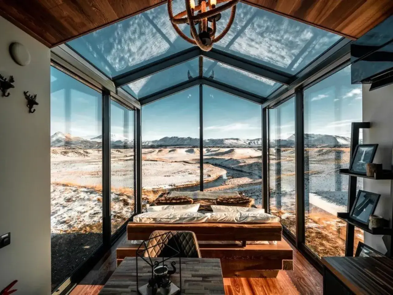 Interior de una casa cápsula con cama y mesa, rodeada de ventanales que ofrecen vistas a un paisaje nevado y montañoso.