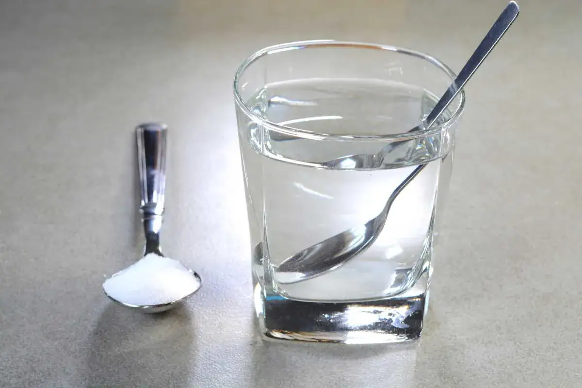 Un bicchiere d'acqua con un cucchiaino e un cucchiaio di sale, un rimedio casalingo per le afte in bocca.
