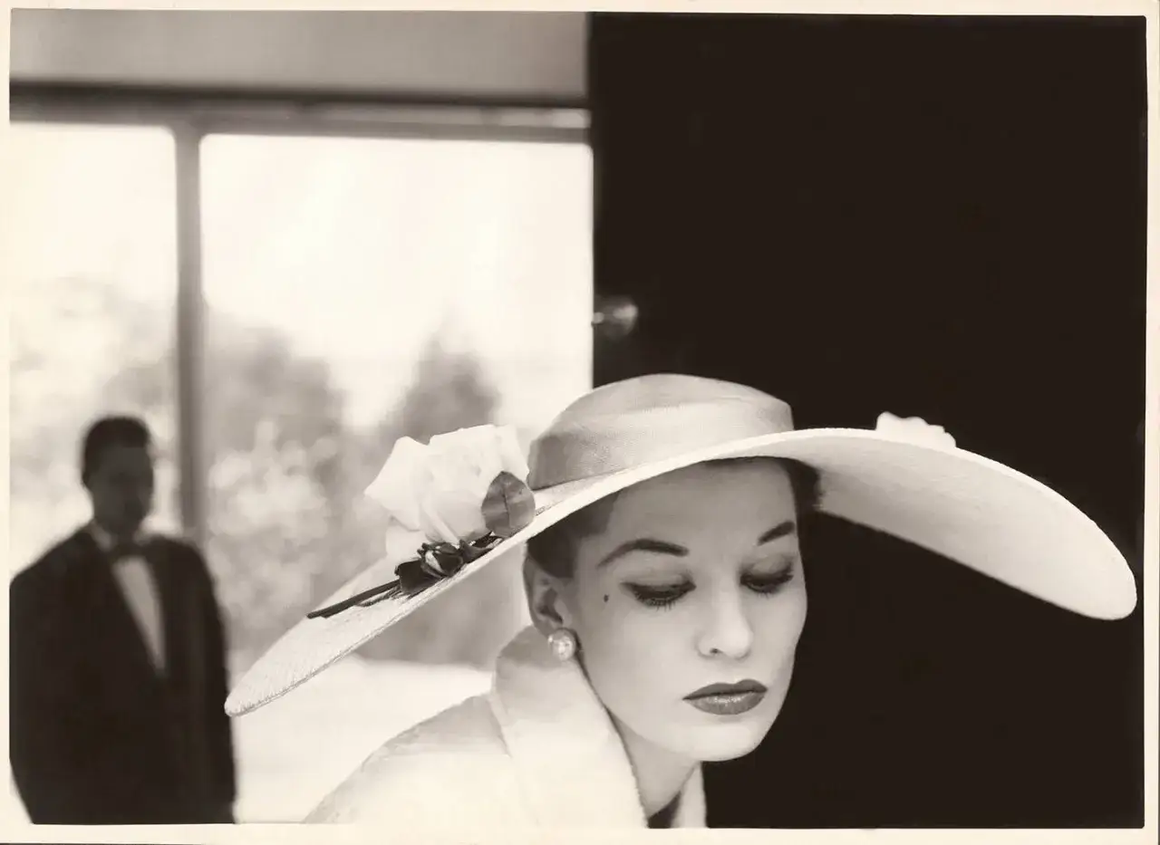 Elegancka kobieta w kapeluszu z różą, w tle mężczyzna w smokingu. Styl Helmut Newton.