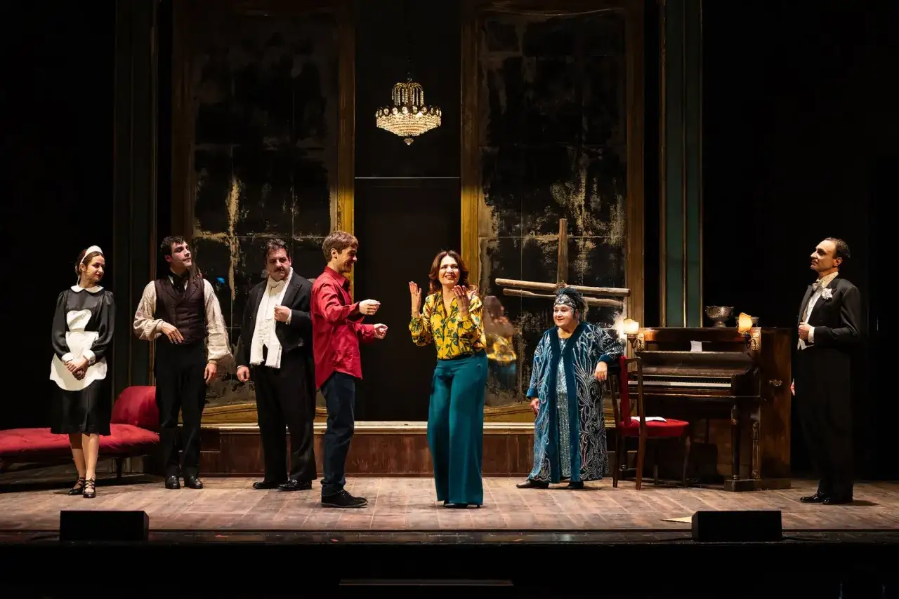 Attori in costume su un palco teatrale. Una donna in abito da cameriera, uomini in abiti eleganti e una donna in un sontuoso abito blu, sembrano un vero animale da palcoscenico.