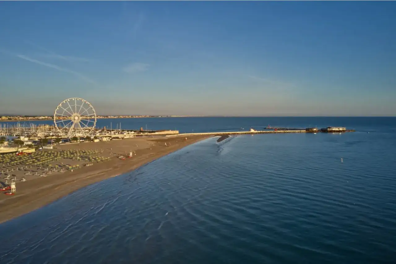 La ruota panoramica domina la spiaggia di Rimini Nord, con il porto e il mare calmo sotto un cielo azzurro.
