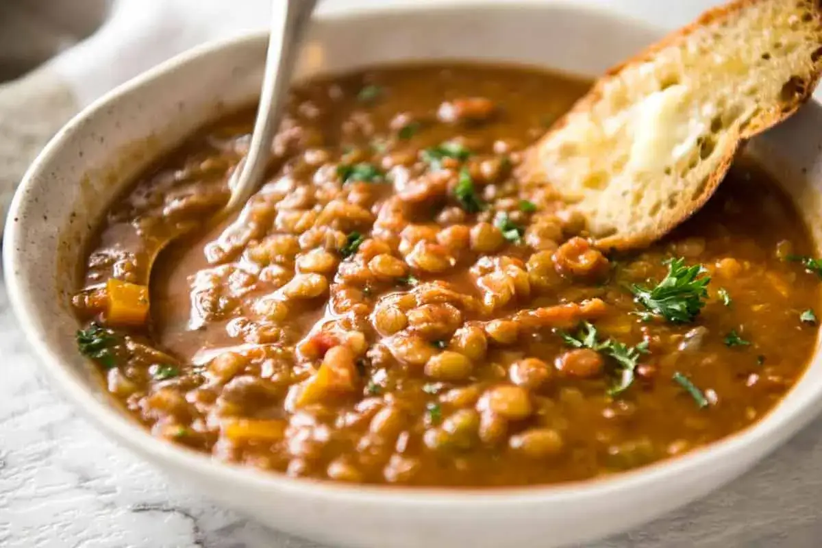 Una ciotola fumante di zuppa di lenticchie, arricchita con verdure e guarnita con prezzemolo fresco, accompagnata da una fetta di pane tostato imburrato.