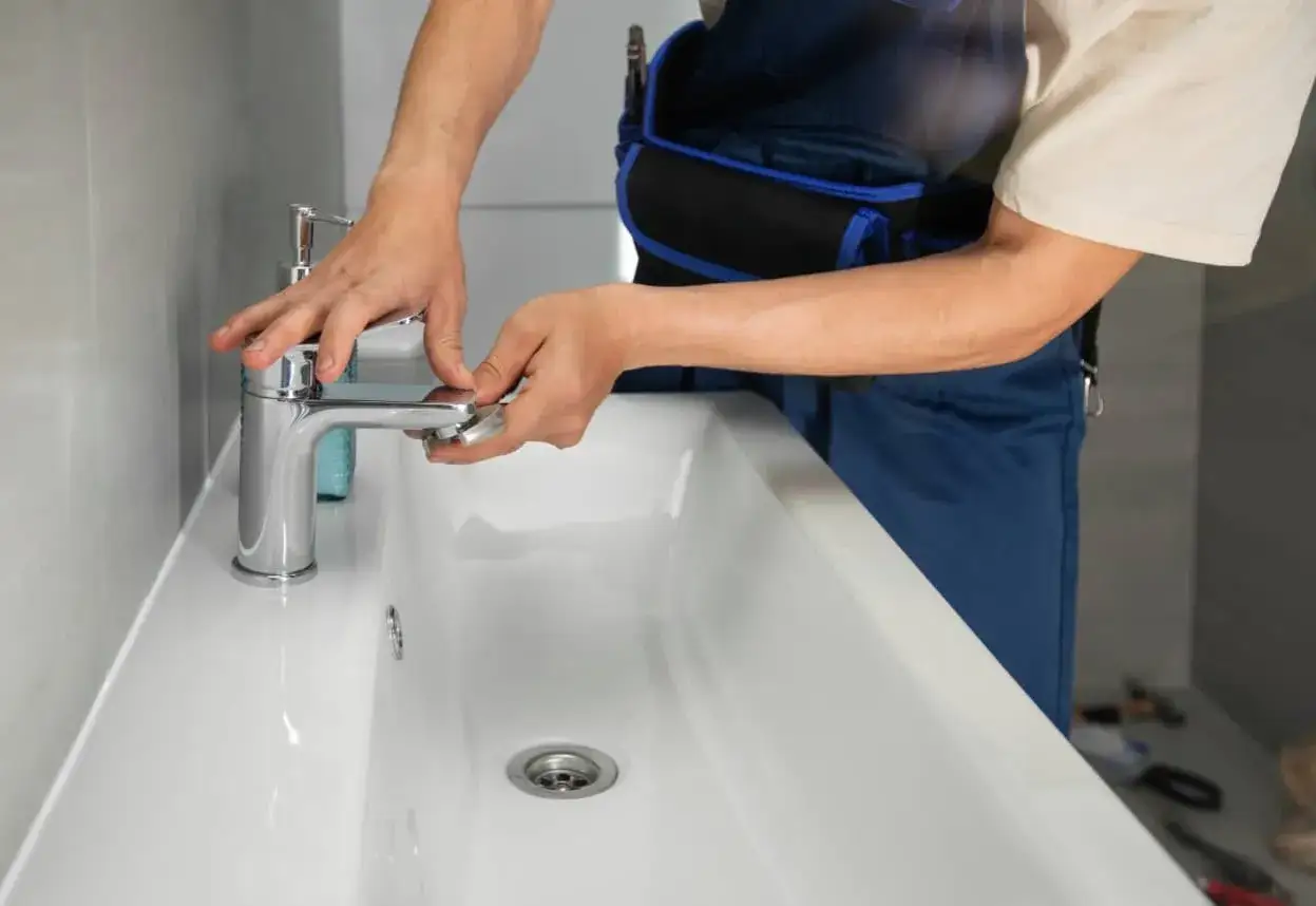Man con mono azul trabajando en un lavabo blanco, ajustando un grifo cromado.