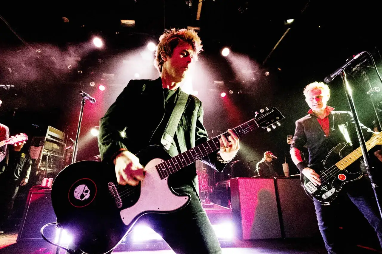 Les membres de Green Day en pleine performance, &eacute;clair&eacute;s par des projecteurs. L'un joue de la guitare, l'autre de la basse.