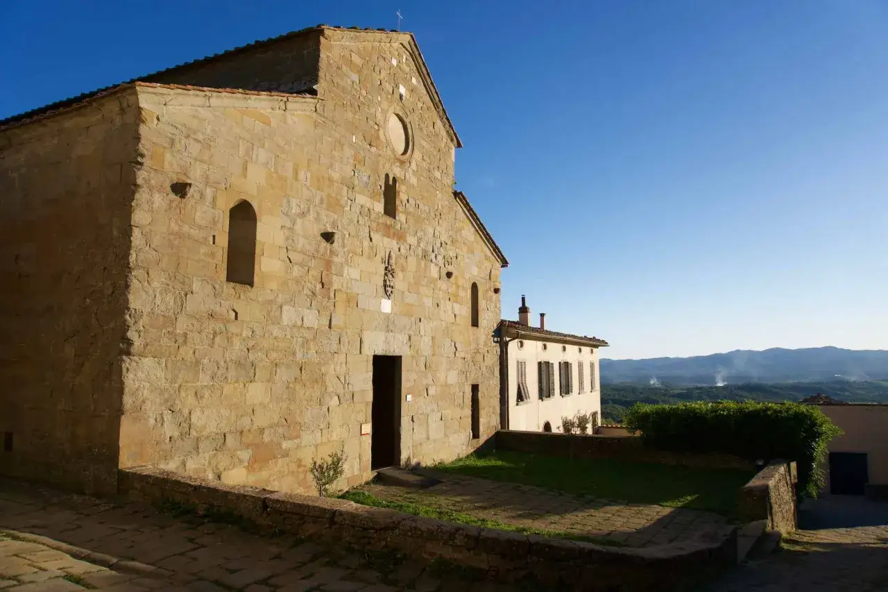 La Pieve di Gropina, con la sua facciata in pietra grezza, si staglia contro un cielo azzurro limpido, affacciata su un paesaggio collinare.