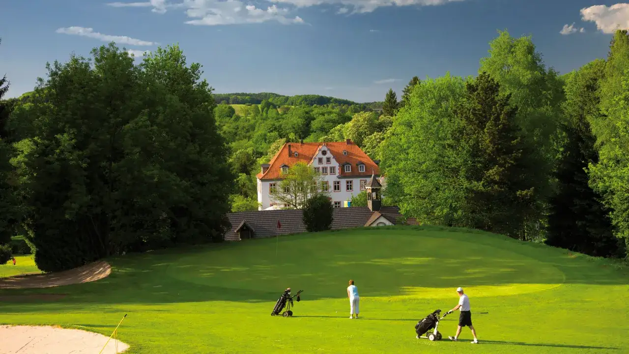 Golfer auf dem grünen Rasen des GC Georghausen, im Hintergrund ein stattliches Gebäude mit rotem Dach, umgeben von üppigem Grün.