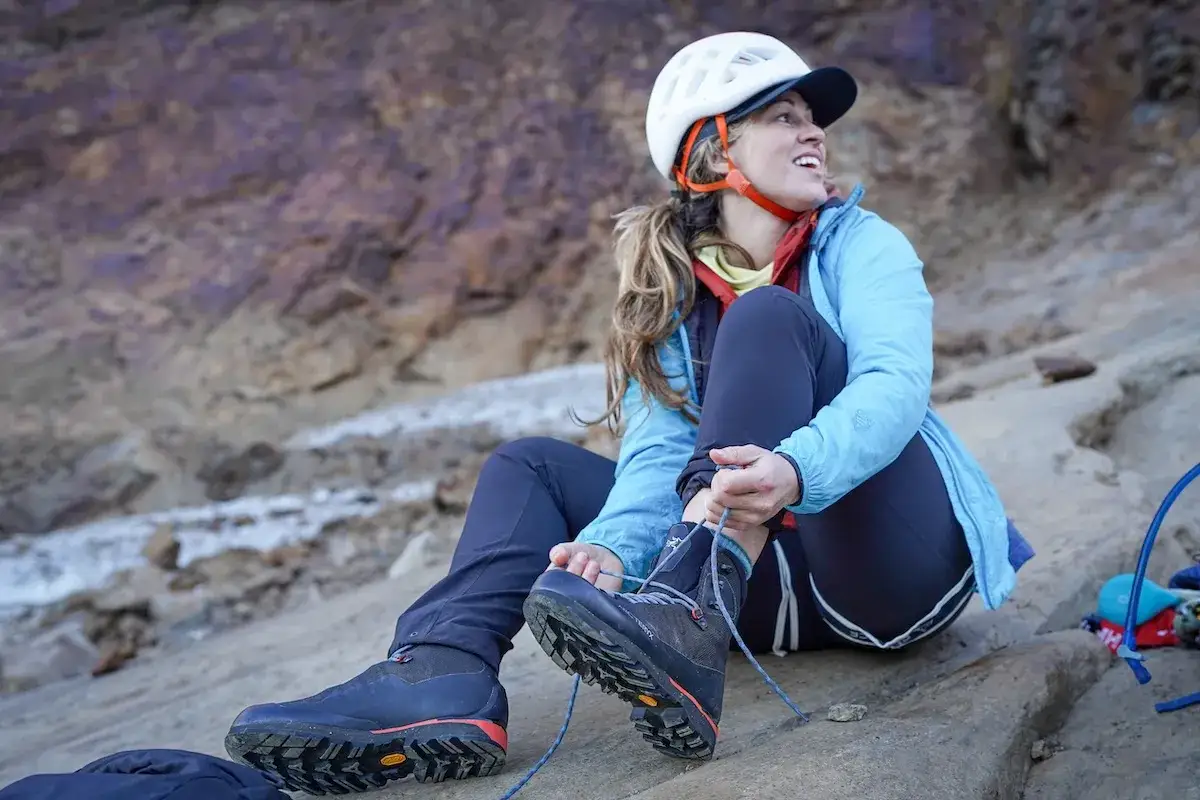A woman in a helmet ties her mountaineering boots vs hiking boots, ready for an adventure on a rocky slope.