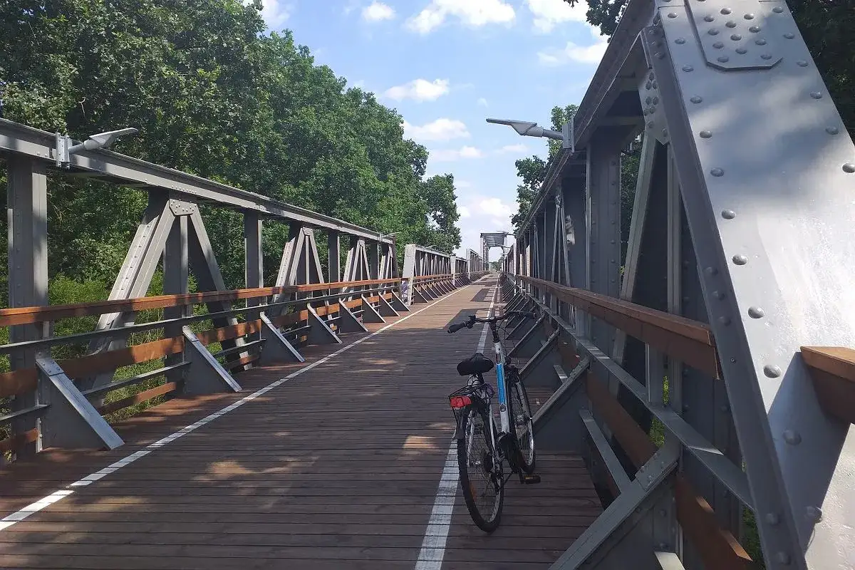 Rower czeka na ścieżce żelazny szlak rowerowy. Drewniany most z metalowymi kratownicami w otoczeniu zieleni.