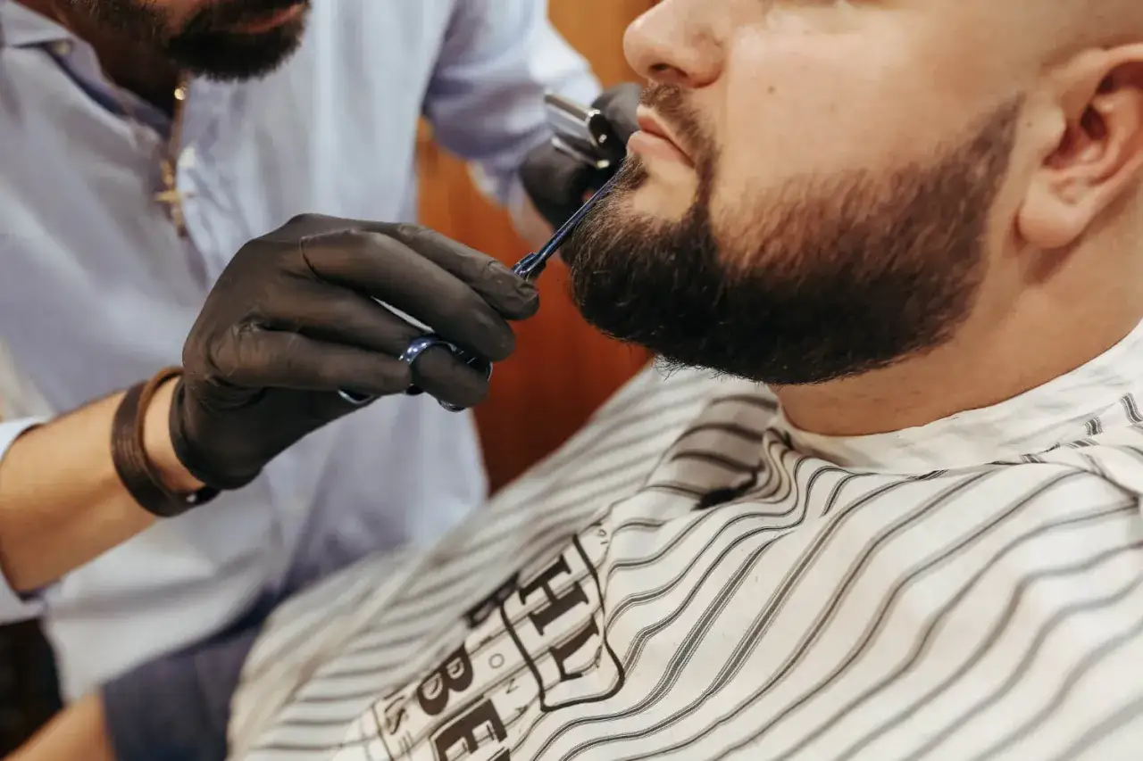 Stylista w czarnych rękawiczkach przycina zarost męski, pokazując jak przyciemnić zarost.