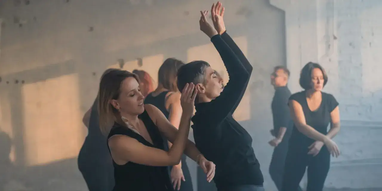 Grupa ludzi w czarnych ubraniach tańczy w zadymionym pomieszczeniu. Jedna z kobiet, kinestetyk, prowadzi ruch.