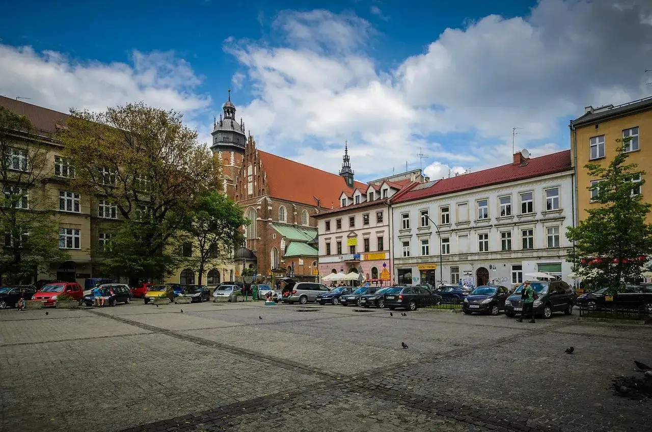 Rynek na części dzielnicy Kazimierz w Krakowie, z kościołem Mariackim w tle i zaparkowanymi samochodami.