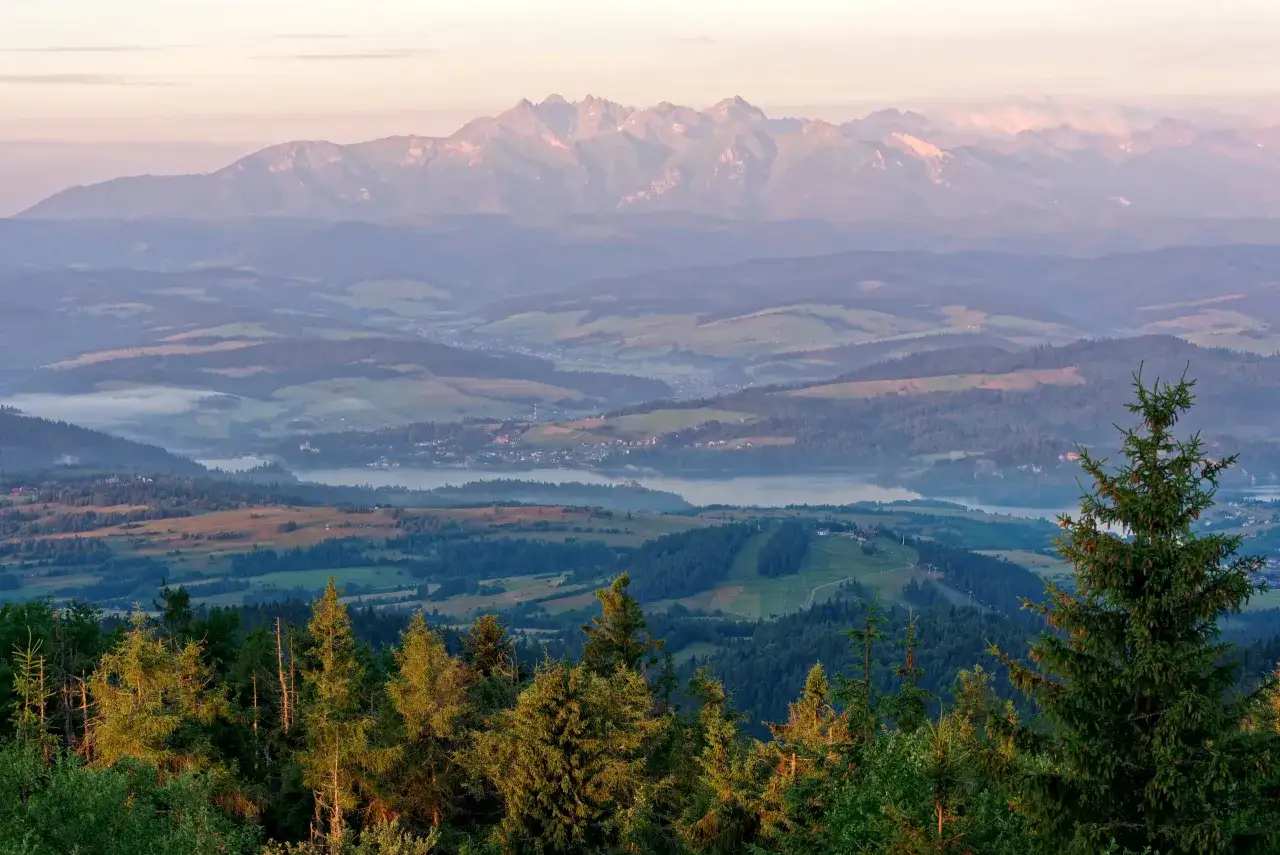 Widok na Tatry o wschodzie słońca. Mgły unoszą się nad doliną, a lasy i pola tworzą malowniczy krajobraz. To miejsce oferuje gorce atrakcje dla miłośników przyrody.