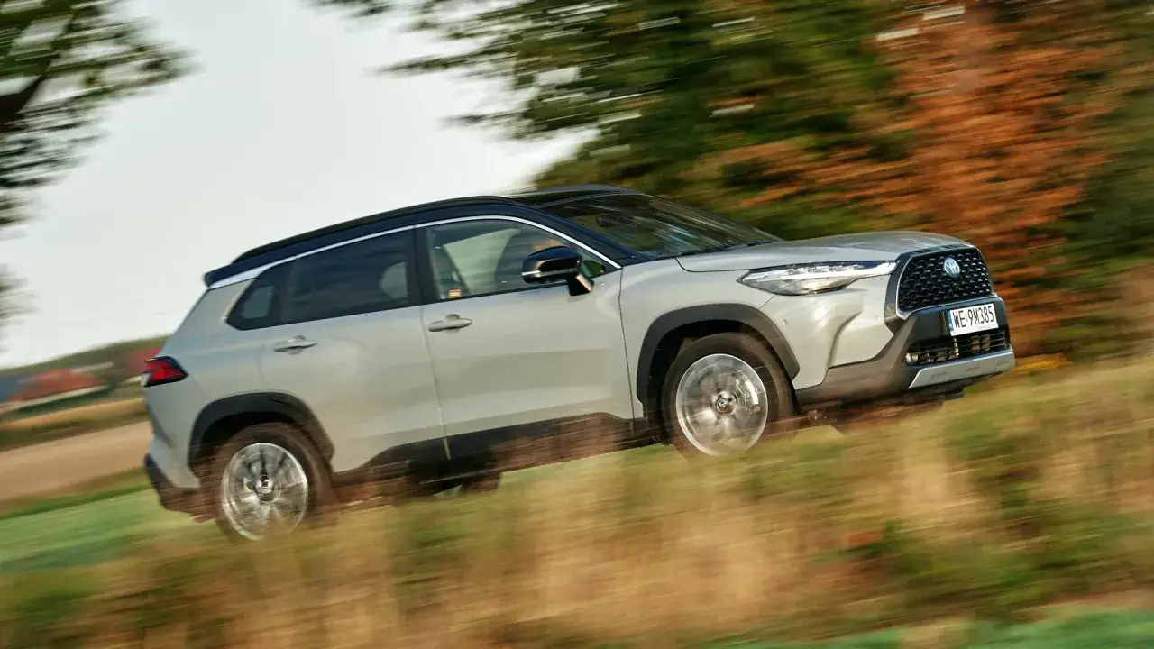 Srebrny Toyota Corolla Cross Hybrid pędzi po wiejskiej drodze, otoczony zielenią i jesiennymi drzewami.