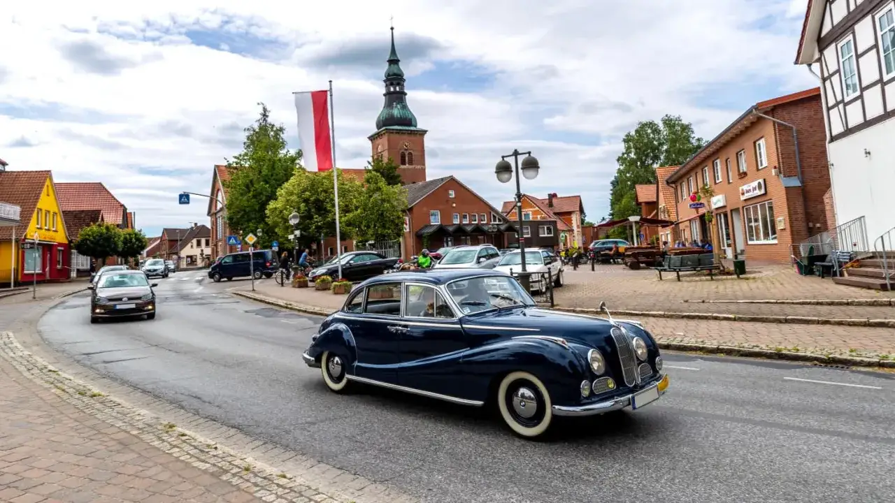 Ein gl&auml;nzender Oldtimer f&auml;hrt durch eine malerische deutsche Stadt. Die Kirche im Hintergrund und die Geb&auml;ude deuten auf eine Umweltzone hin, in der solche Klassiker willkommen sind.