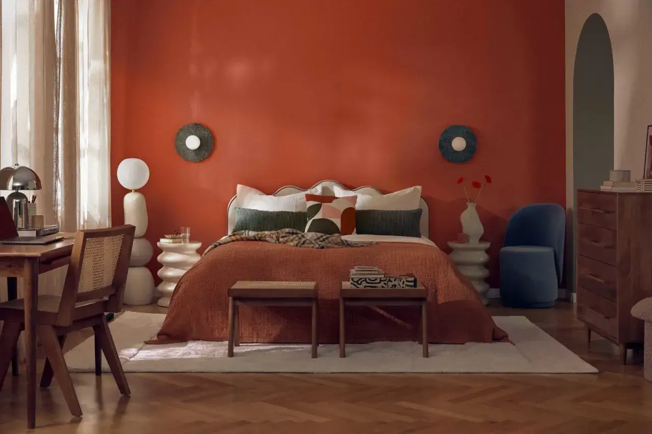 Dormitorio con pared naranja, cama con colcha terracota y cojines geométricos. Destacan las tendencias interiorismo con muebles de madera y detalles curvos.