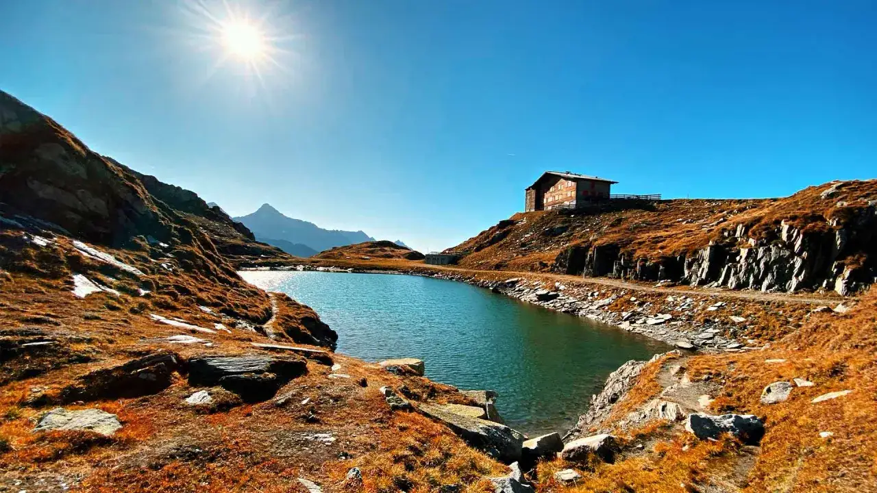 Ein Bergsee glitzert unter strahlender Sonne. Am Ufer thront das Pfitscher Joch Haus, umgeben von herbstlich gef&auml;rbten H&auml;ngen und Felsen.
